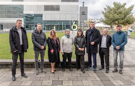 Acht Personen – vier Frauen und vier Männer – in herbstlicher Business-Casual-Kleidung posieren nebeneinander auf einem gepflasterten Platz. Im Hintergrund sind eine Rasenfläche, ein modernes Glasgebäude mit der Ziffer 6 und ein Schild mit der Aufschrift „platz der vielfalt“ zu sehen.